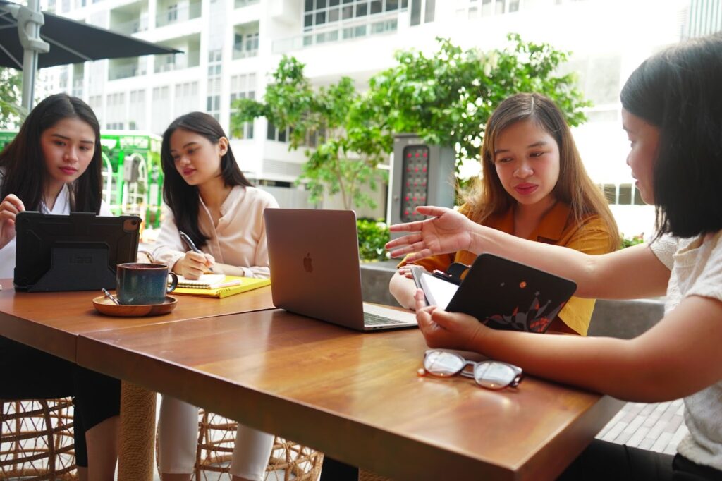 Four women are collaborating on a business project outdoors, using laptops and tablets, and discussing forward planning strategies for future growth.